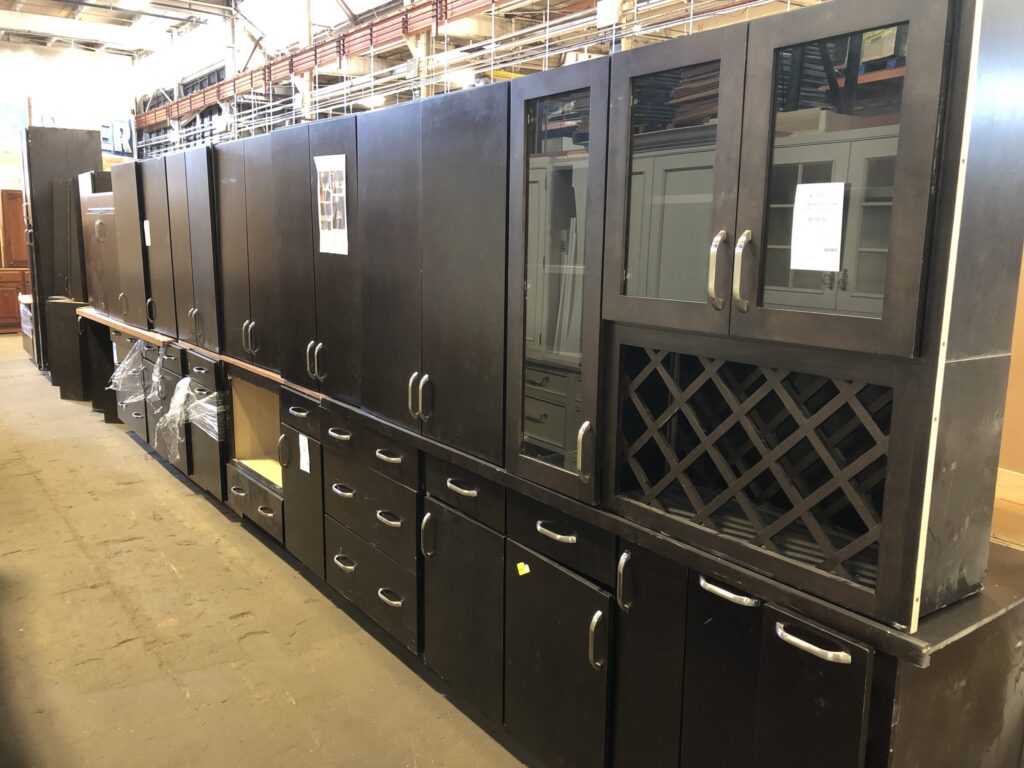 Long row of dark brown salvaged kitchen cabinets with silver hardware, some featuring glass fronts and an integrated wine rack, available at the Baltimore non-profit thrift store, offering affordable used building materials for sustainable home improvement.