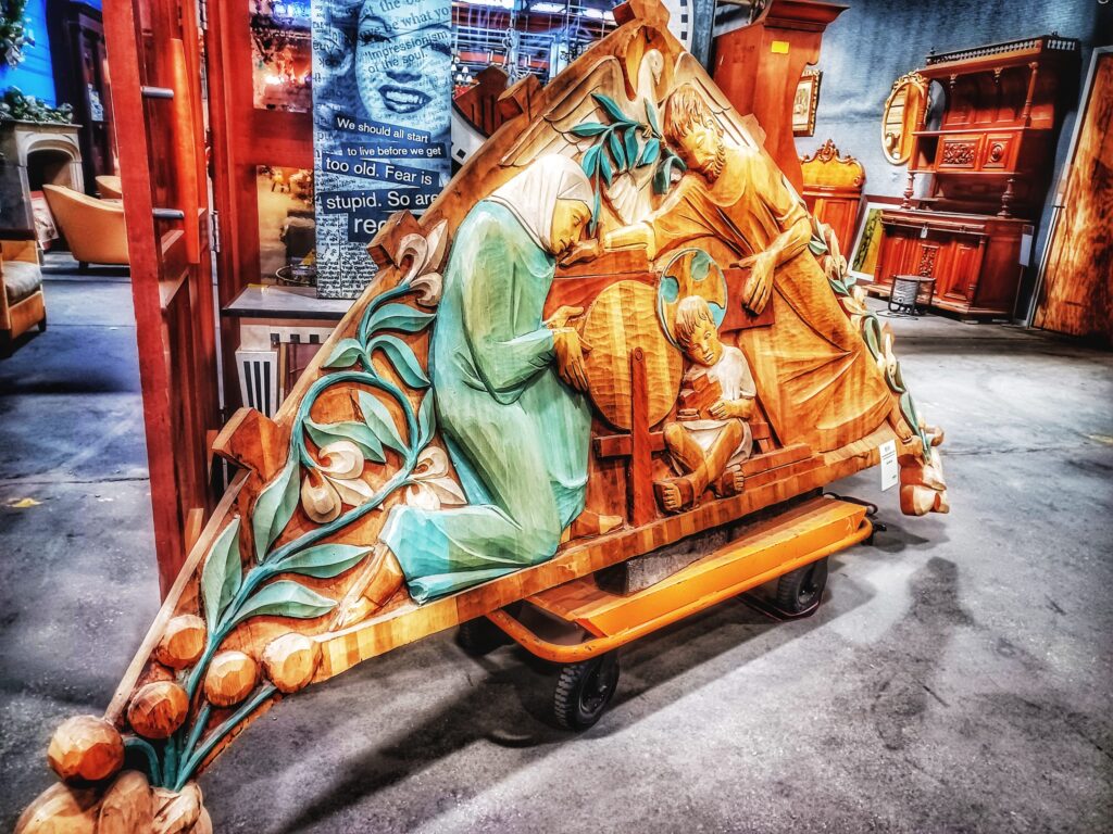 Large, intricately carved salvaged wood relief with teal painted accents and figures, displayed on a cart at the Baltimore non-profit Second Chance. Background shows vintage furniture and other unique secondhand building materials and home decor.