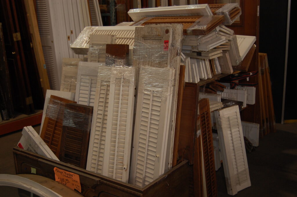 Large stack of salvaged and secondhand building materials including white louvered shutters, vintage wood doors, and antique window shutters available for home improvement and unique decor at the Baltimore non-profit, Second Chance.